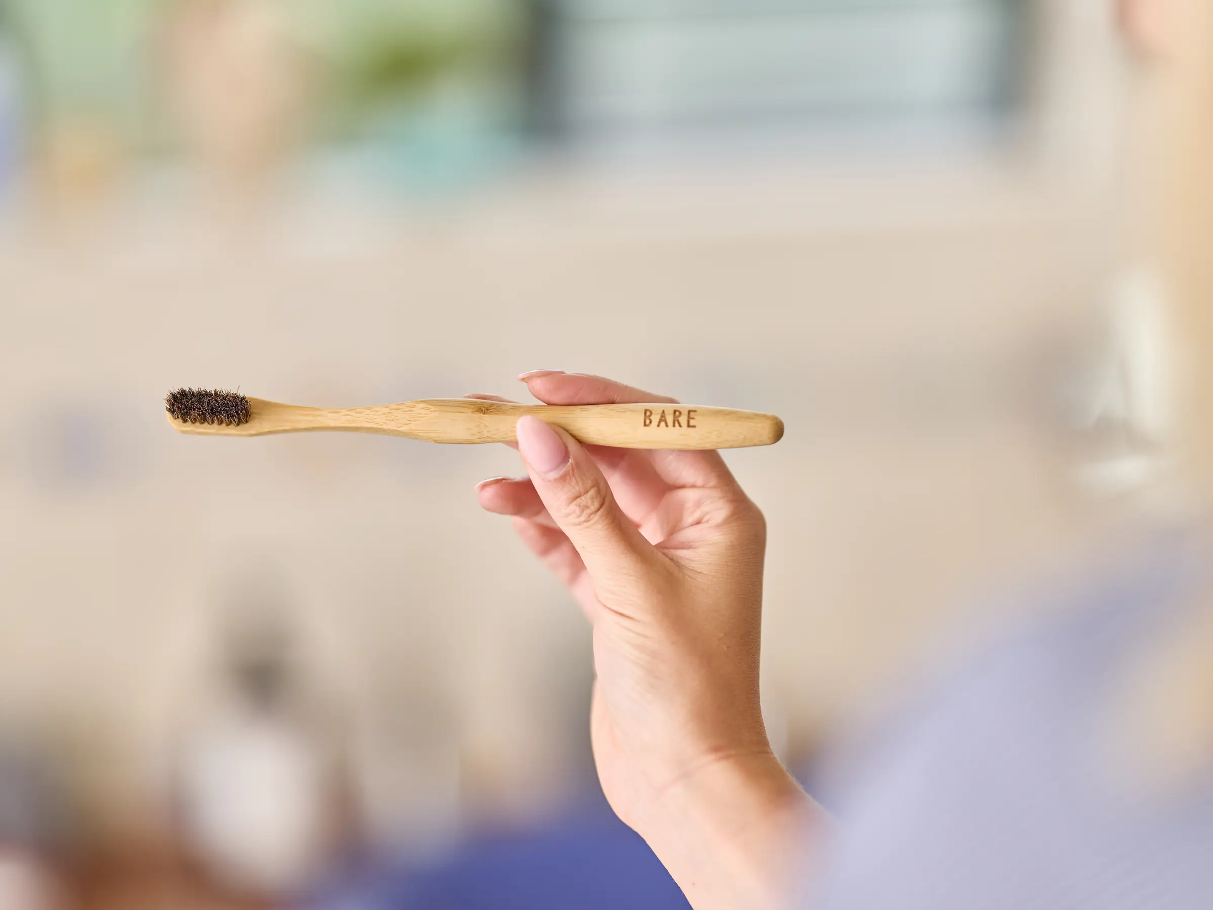 A hand holding the BARE toothbrush level in a sunlit bathroom, the BARE engraving on the bamboo handle and the dark horse-hair bristles in focus