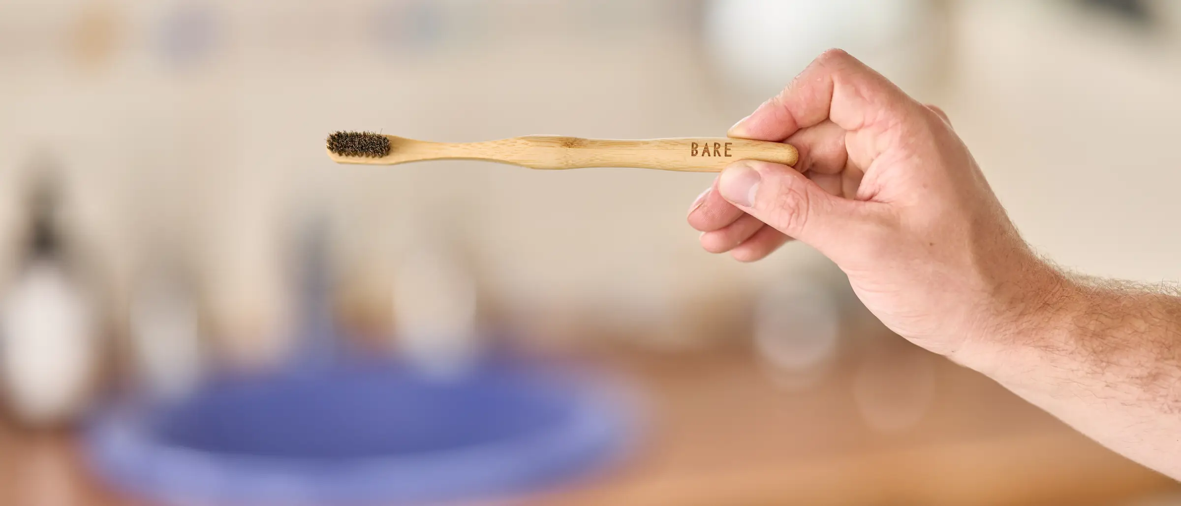 A hand holding the BARE toothbrush over a timber bathroom vanity, a blue basin soft in the background