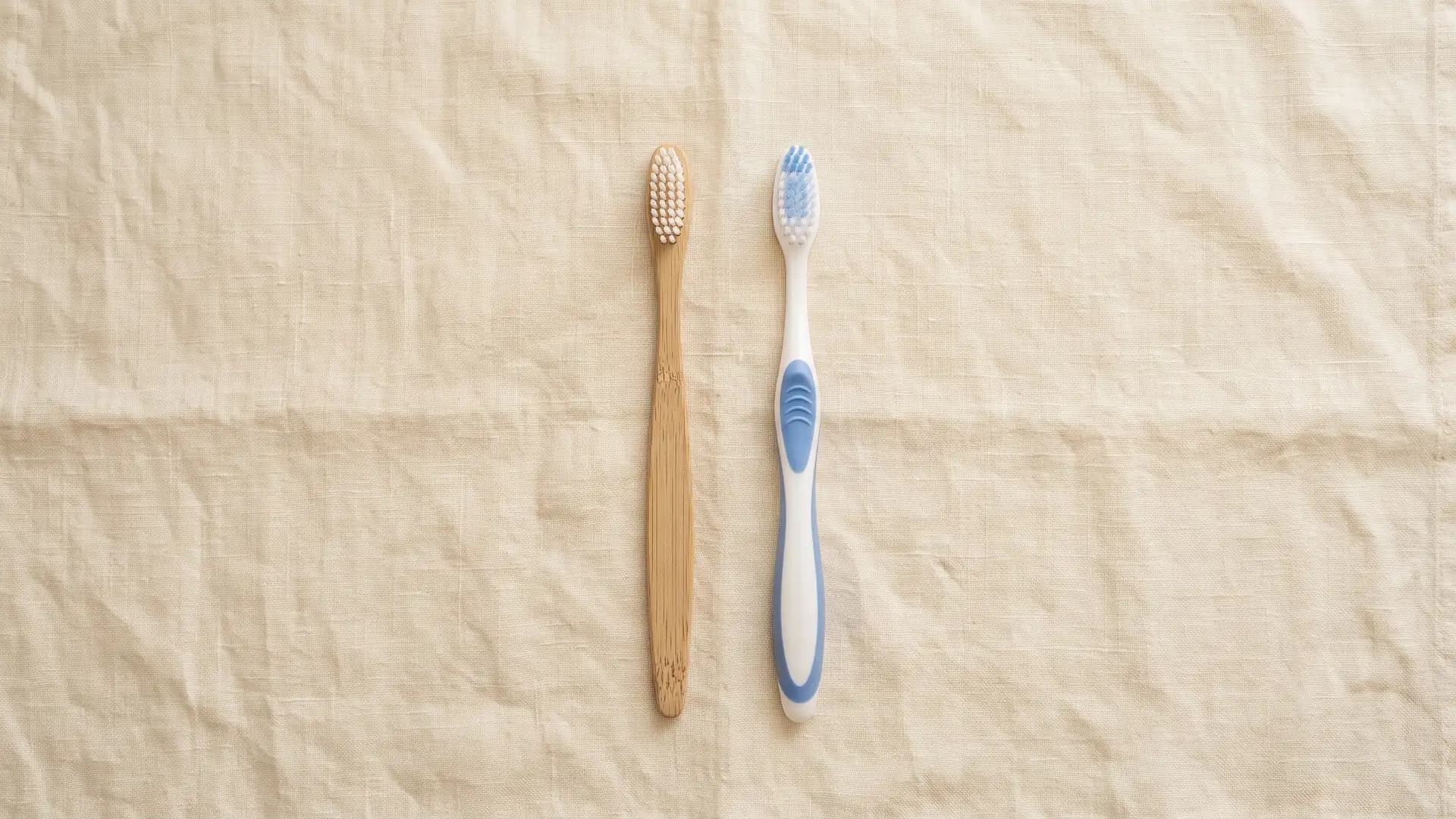 A bamboo toothbrush and a white plastic toothbrush with a blue grip lying side by side on cream linen, photographed from above.