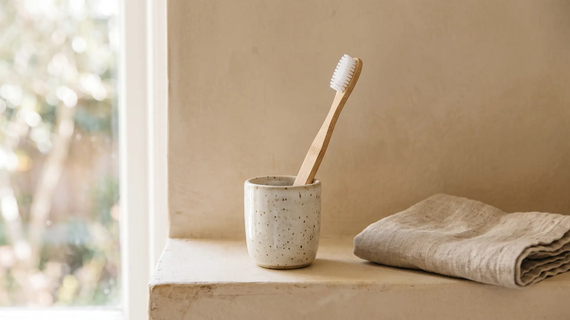 An ordinary bamboo toothbrush with white nylon bristles standing in a speckled ceramic cup on a bathroom shelf beside a folded linen towel, in soft window light.