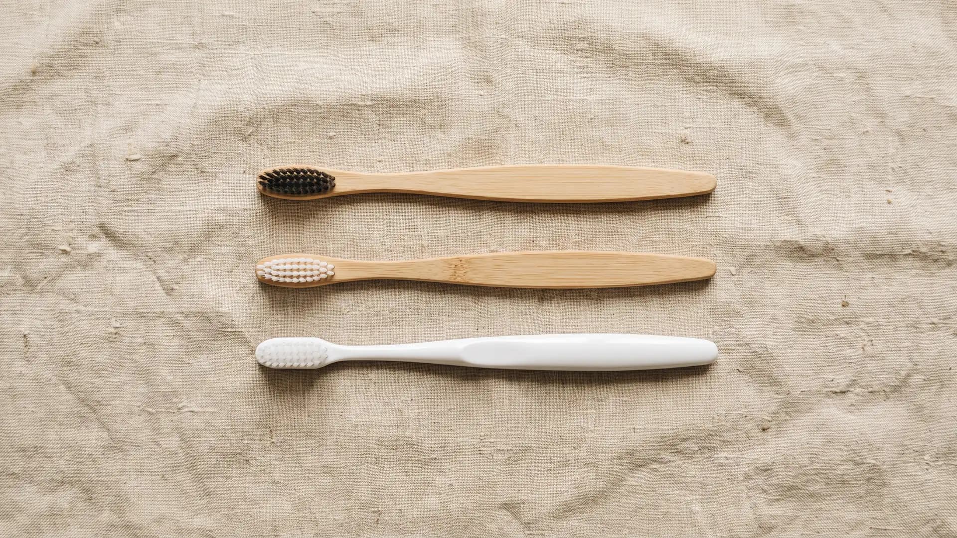 Three toothbrushes on cream linen — a white plastic brush, a bamboo brush with white nylon bristles, and a bamboo brush with dark natural bristles — the three material recipes this page takes apart.