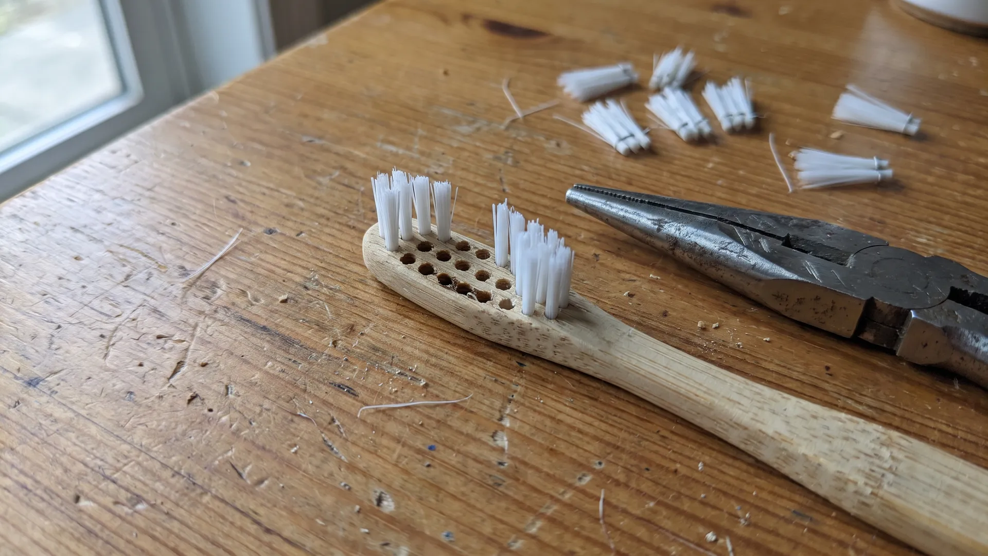 A bamboo toothbrush head part-way through de-bristling, with several tufts pulled out leaving empty holes in the wood and needle-nose pliers alongside.